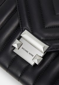 Musta nahkalaukku, jossa on tyylikäs tikattu muotoilu, hopeiset yksityiskohdat ja näkyvä solki, jossa on brändin nimi "Michael Kors."