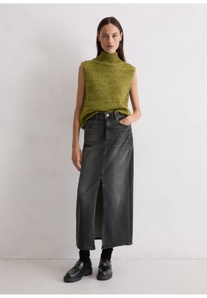 Pull à col roulé sans manches vert et jupe maxi en denim noir avec fente frontale. La mannequin se tient avec la main dans la poche de la jupe, chaussures noires.