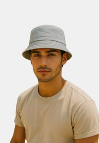 Cappello a secchiello grigio realizzato in tessuto morbido con una larga visiera, abbinato a una maglietta beige semplice. La texture appare liscia, senza motivi visibili.