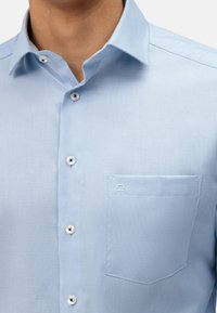 Světlomodrá košile s knoflíky, texturovaná tkanina, špičatým límečkem a levou náprsní kapsou s malým logem.