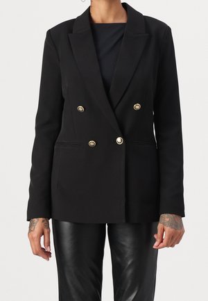 Osoba ubrana w czarną, dwurzędową marynarkę z złotymi guzikami i czarne skórzane spodnie, z widocznymi tatuażami na dłoniach.