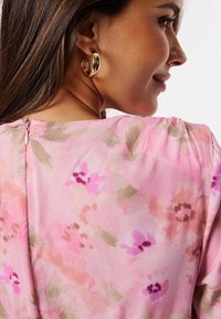 Blus med blommönster i ljusrosa tyg. Har en nyckelhålsrygg med dragkedja och en mjuk, flödande textur. Accenterad med guldörhängen i ring. 