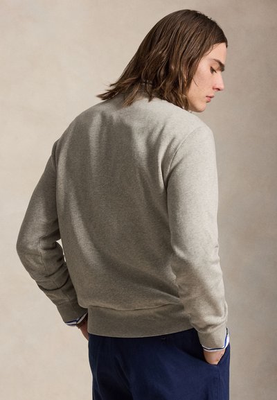 Jeune homme aux cheveux bruns mi-longs, portant un sweat-shirt gris clair et un pantalon bleu marine, debout les mains dans les poches, de profil.