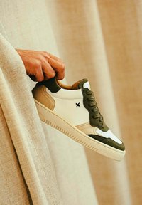 Une personne tenant une sneaker blanche et vert olive avec des accents beiges sur un fond en tissu neutre.