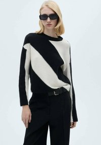 Pull en tricot à rayures diagonales noir et blanc avec un col rond, présentant des manches longues et une coupe décontractée. Associé à un pantalon noir.