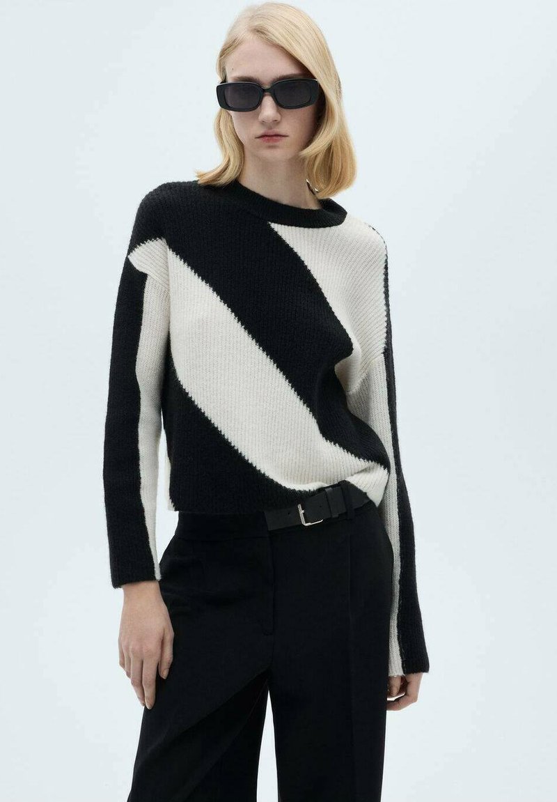 Pull en tricot à rayures diagonales noir et blanc avec un col rond, présentant des manches longues et une coupe décontractée. Associé à un pantalon noir.