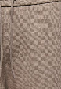 Gros plan sur un pantalon en tissu beige avec un motif en armure diagonale et un cordon suspendu à côté de l'ouverture d'une poche.