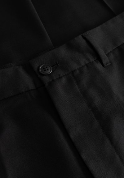 Sort stof med en glat tekstur, der har lukning med knapper, bæltestropper og et skræddersyet design. Syningerne er tydeligt synlige.