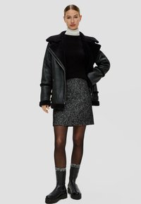 Veste en cuir noir avec doublure en fourrure, pull noir à côtes, jupe texturée grise, collants noirs et bottines noires avec des poignets à motifs.
