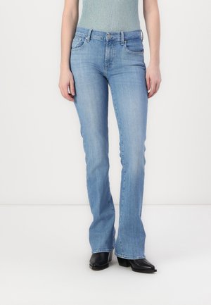 Femme portant un jean bootcut bleu clair, des bottes noires à bout pointu, et un haut sans manches texturé sur un fond blanc.