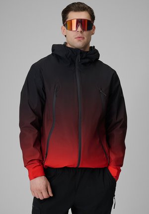 Mężczyzna w czarno-czerwonej gradientowej kurtce na zamek z kapturem i czerwonych lustrzanych sportowych okularach przeciwsłonecznych na szarym tle.