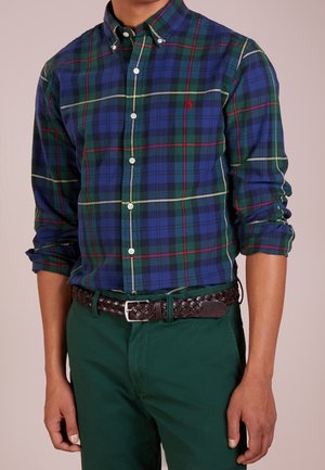 Homme portant une chemise à carreaux bleue, verte et rouge à manches retroussées, un pantalon vert foncé et une ceinture en cuir tressé marron.