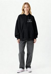 Fekete, oversize kapucnis pulóver, húzózsinórral és elülső zsebbel, amelyen a fehér "Hôtel de L'Amour" felirat található. Laza, sötétszürke farmernadrággal és fehér sportcipővel párosítva.