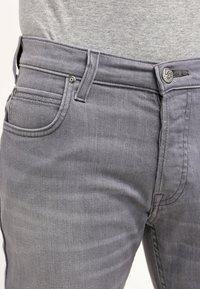 Jeans de mezclilla gris con un diseño ajustado, estilo clásico de cinco bolsillos y un botón metálico grabado con "Lee". Textura suave.