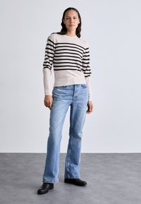 Pull rayé beige et noir, associé à un jean évasé bleu clair. Le modèle porte des bottines à talons noires. Textures lisses, style décontracté.