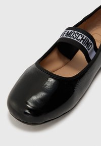 Črne balerinke z sijajno lakasto površino, elastičnim trakom z belim logotipom in obliko zaokroženega prsta. Gladka notranja podloga.