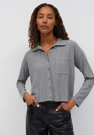 Vrouw met krullend haar die een grijs geribd blouse met knopen en lange mouwen draagt en zwarte leren broek met handen in de zakken.