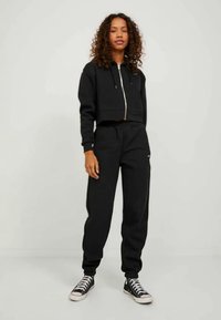 Corta-vento preto com fecho éclair e calças de treino combinando, feitas de tecido suave, com cordão na cintura e punhos ajustados, combinadas com ténis pretos.