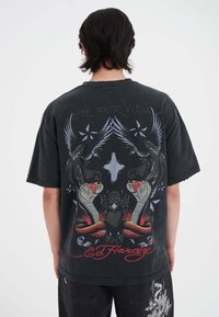 Sort oversized t-shirt med grafik af to slanger og fugle, med teksten "Arte Por Vida" og "Ed Hardy" i fed rød. Slidte kanter.