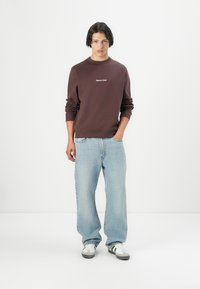 Mørkebrun sweatshirt med rund hals og hvit brodert logo, kombinert med lyseblå vid leg jeans og hvite sneakers med svarte detaljer.