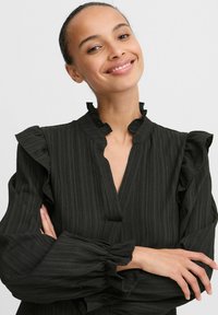Blusa nera con righe verticali, collo e polsini arricciati. Presenta maniche lunghe e una vestibilità rilassata. Texture liscia, design minimalista.
