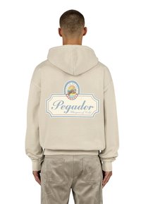 Felpa beige con cappuccio e cordino, caratterizzata da una grande grafica stampata sul retro che include testo e un design paesaggistico.