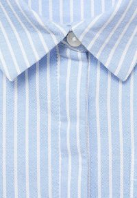 Material în dungi albastre deschise și albe, la o privire de aproape, având un nasture la guler și o țesătură texturată. Dungile sunt distanțate uniform.