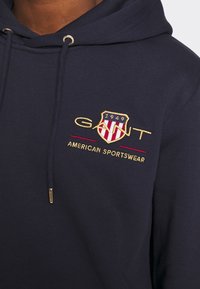 Sudadera azul marino con logotipo y texto bordados en oro y rojo. Cuenta con cordones y una tela suave y texturizada.