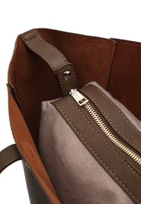 Brązowa skórzana torba typu tote z miękkim zamszowym wnętrzem oraz wewnętrzną kieszenią zamykaną na zamek z jasnoszarego materiału, zbliżenie na zamek i szycie.