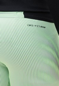 Κοντινό πλάνο σε αθλητικό κολάν με ρίγες σε νέον πράσινο και μαύρο χρώμα με το λογότυπο "DRI-FIT ADV" στο δεξί ισχίο, σε συνδυασμό με μαύρο τοπ.