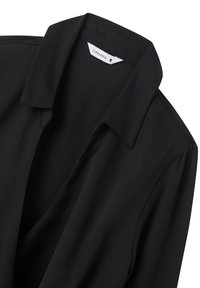 Blusa nera con colletto, texture liscia e design drappeggiato. L'etichetta mostra la taglia S. Il tessuto sembra leggero ed elegante.