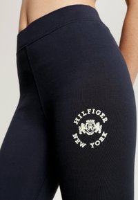 Granatowe legginsy wykonane z elastycznego materiału, z białym okrągłym logo z napisem "HILFIGER NEW YORK" oraz dekoracyjnym herbem na udzie.