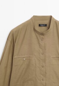 Khaki-shirt met een mandarin-kraag, knoopsluiting aan de voorkant en twee voorzakken. Gemaakt van lichtgewicht stof met een gladde textuur en subtiele glans.