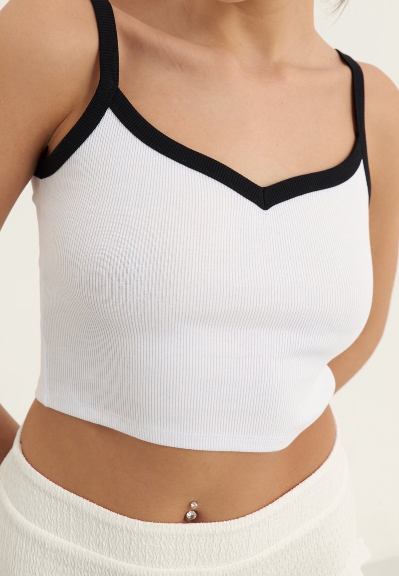 Top corto blanco de canalé con un ribete en contraste negro y un diseño de escote en V. Presenta tirantes finos y una silueta ajustada.