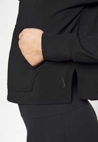 Schwarzes Langarmtop mit einer vorderen Tasche, abgerundetem Saum und dezentem Nike-Logo in passendem Farbton. Glatter, dehnbarer Stoff.