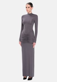 Mujer de pie, vistiendo un vestido gris ajustado de manga larga con detalles fruncidos en la cintura y un escote alto, combinado con tacones negros.