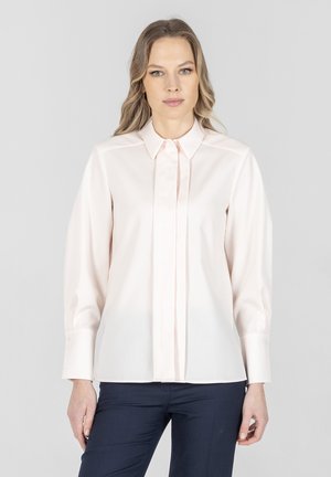 Kobieta o długich blond włosach, ubrana w jasnoróżową bluzkę z guzikami i ciemnoniebieskie spodnie, stojąca na tle jednolitego, szarego tła.