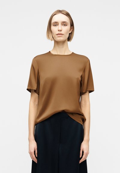 Γυναίκα με ξανθά κοντά μαλλιά φοράει καφέ κοντομάνικο μπλουζάκι και μαύρο παντελόνι, στέκεται μπροστά σε απλό φωτεινό φόντο.