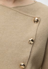 Vêtement en laine beige avec un décolleté asymétrique maintenu par trois boutons ronds dorés brillants au niveau de l'épaule.