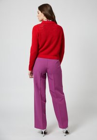 Maglione rosso lavorato a maglia con un motivo testurizzato, abbinato a pantaloni a vita alta e gamba larga in viola. Tacchi a spillo bianchi visibili.
