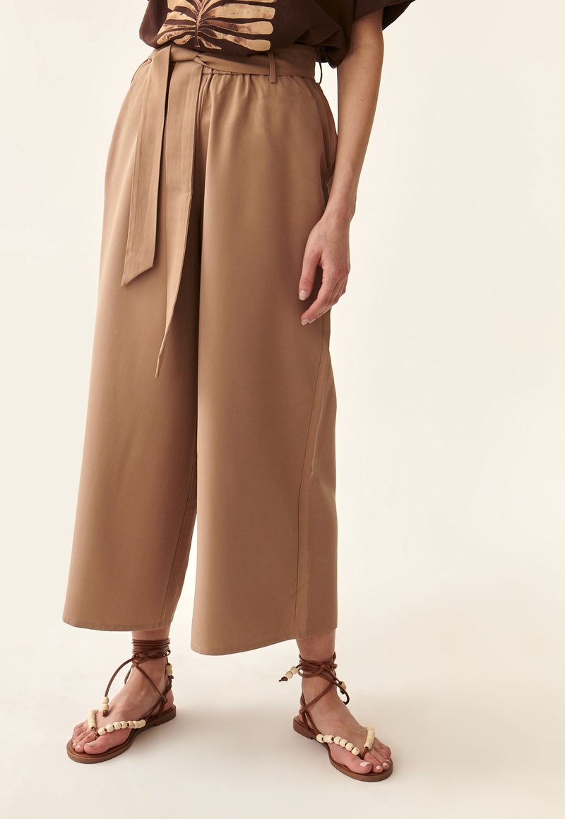 Personne portant un pantalon beige à jambes larges avec une ceinture en tissu, un haut marron imprimé et des sandales marron à brides perlées blanches.