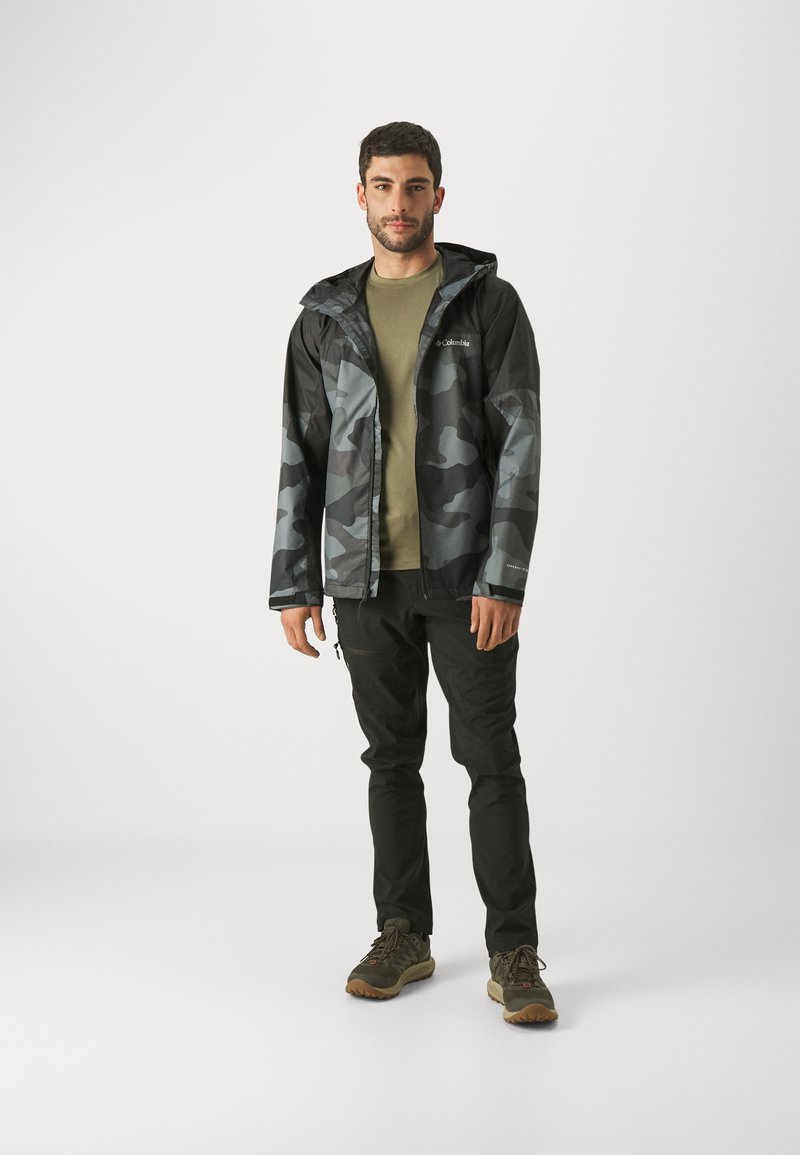 Chaqueta de camuflaje negra y gris resistente al agua con capucha, llevada sobre una camiseta verde oliva, combinada con pantalones negros y zapatos de exterior.