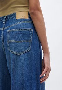 Τζιν παντελόνια με φαρδιά μπατζάκια σε βαθύ μπλε χρώμα, με δερμάτινη ταμπέλα ταμπά στη μέση και πίσω τσέπη με κίτρινη ραφή λεπτομέρειες.