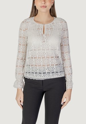 Blusa bianca in pizzo con motivo floreale, scollatura rotonda, dettaglio a occhiello, maniche lunghe e orlo scallop, abbinata a pantaloni neri.