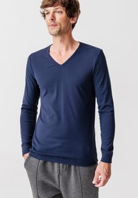Maglietta a maniche lunghe a V di colore navy in tessuto morbido, con un design aderente e una texture liscia. Indossata con pantaloni grigi con cordino.