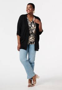 Zwarte open voorkardigan over een gebloemde blouse, lichtblauwe rechte jeans en beige sandaaltjes met bandjes. Gestructureerde stof en opgerolde mouwen.