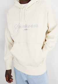 Krémová mikina s kapucí, na které je vyšívaný nápis "Sportswear by PUMA" světle modrou barvou. Obsahuje ribbené manžety, klokánkovou kapsu a jemnou strukturu.