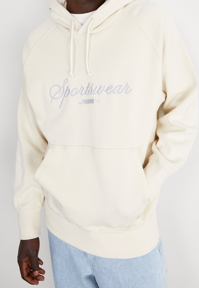 Felpa con cappuccio color crema, con "Sportswear by PUMA" ricamato in azzurro. Presenta polsini a coste, tasca a marsupio e una texture morbida.