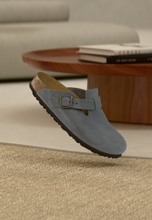 Un único zapato gris de ante sin cordones con hebilla, plantilla de corcho y suela negra flotando sobre una alfombra beige en un salón.
