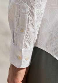 Witte geborduurde blouse met lange mouwen, versierd met bloemenpatronen, zichtbare stiksels en twee ronde knopen op de manchetten. Glad textuur.